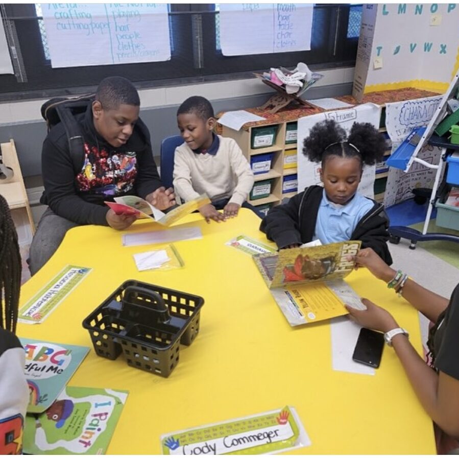 Older Student Reading with Younger Peers in Classroom A group of students and an adult sit around a bright yellow table in a classroom, reading books together. A teen is reading to a younger child, while another child listens and reads with a teacher. The setting includes colorful books, name tags, and educational posters on the wall.
