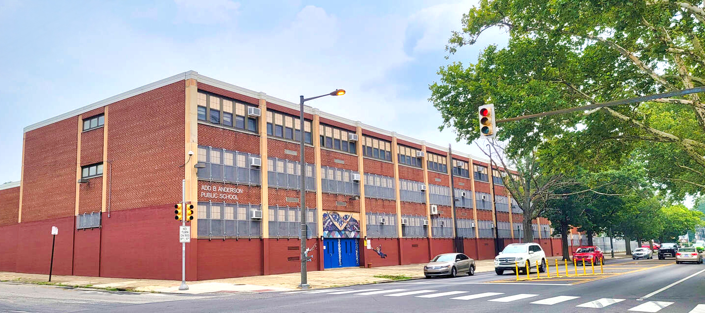 Add B. Anderson Public School Building Exterior Exterior view of Add B. Anderson Public School, a large red-brick school building with multiple windows and a blue front entrance, located on a city street lined with trees and cars.