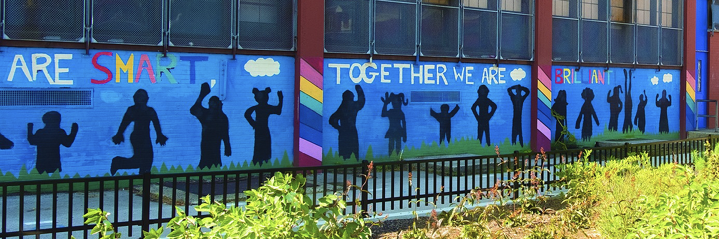 Mural on a school building with colorful text that reads “ARE SMART TOGETHER WE ARE BRILLIANT,” featuring black silhouettes of children in playful poses against a bright blue background with clouds, grass, and rainbow stripes.