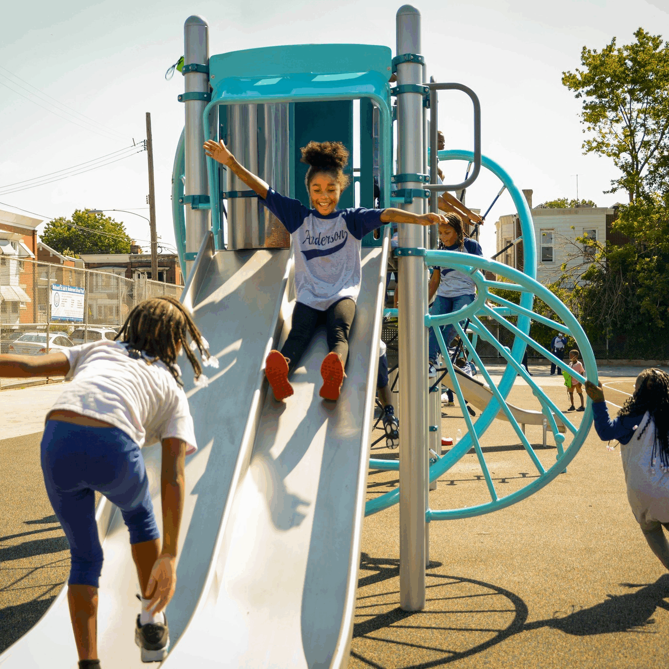 Add B. Anderson students playing on the school playground slide Young students playing on a bright blue and silver playground slide at Add B. Anderson School. One child is sliding down joyfully with arms outstretched while others climb and play around the structure on a sunny day.